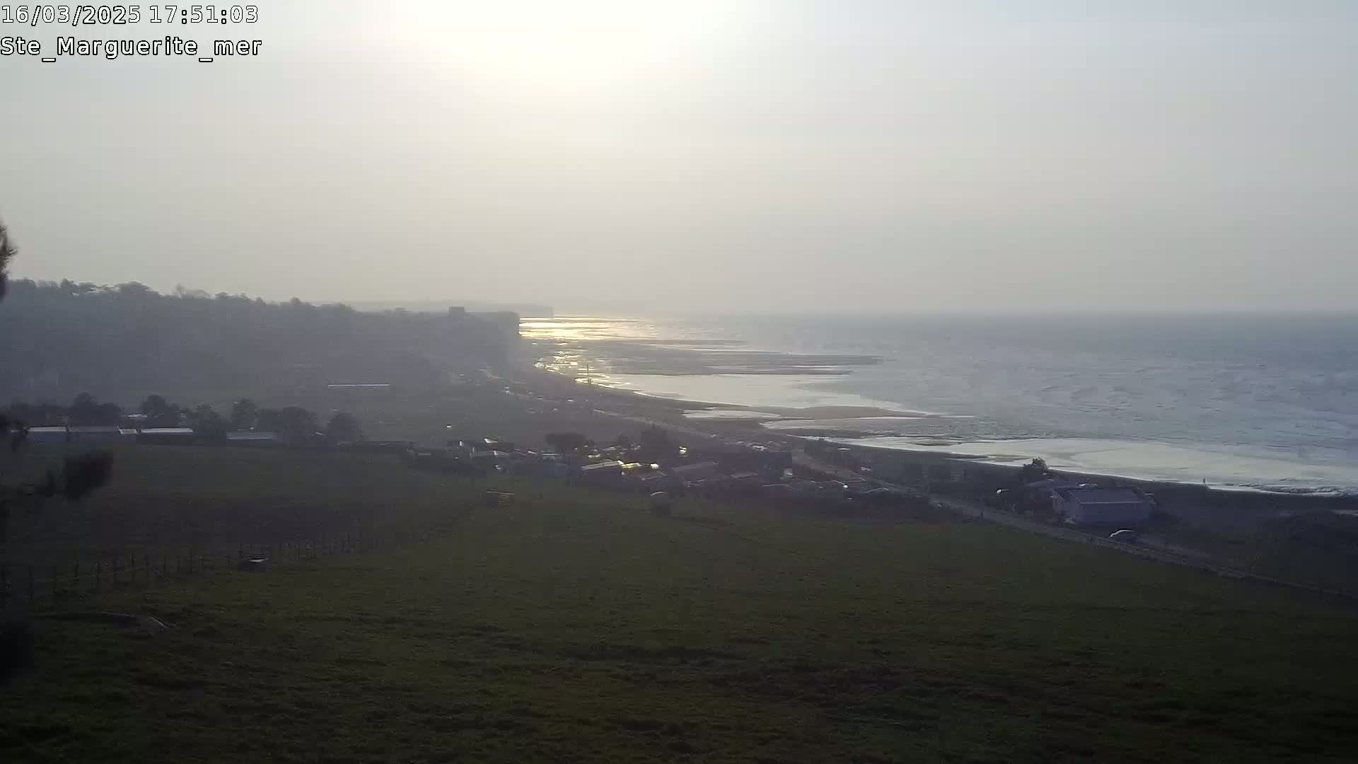 A hazy sunset illuminates a coastline with a village and grassy fields visible from a high vantage point.