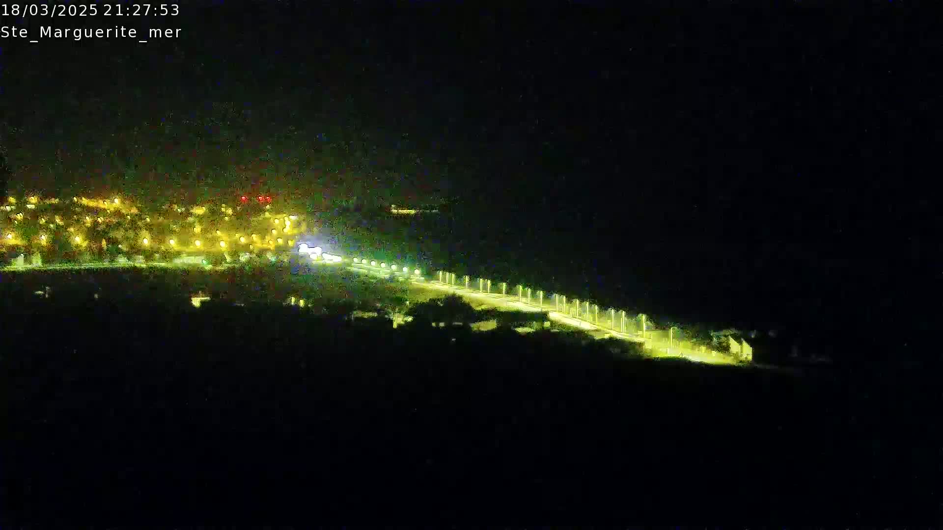 A nighttime, long-range view of a brightly lit roadway stretching from a cluster of illuminated buildings toward a dark body of water.