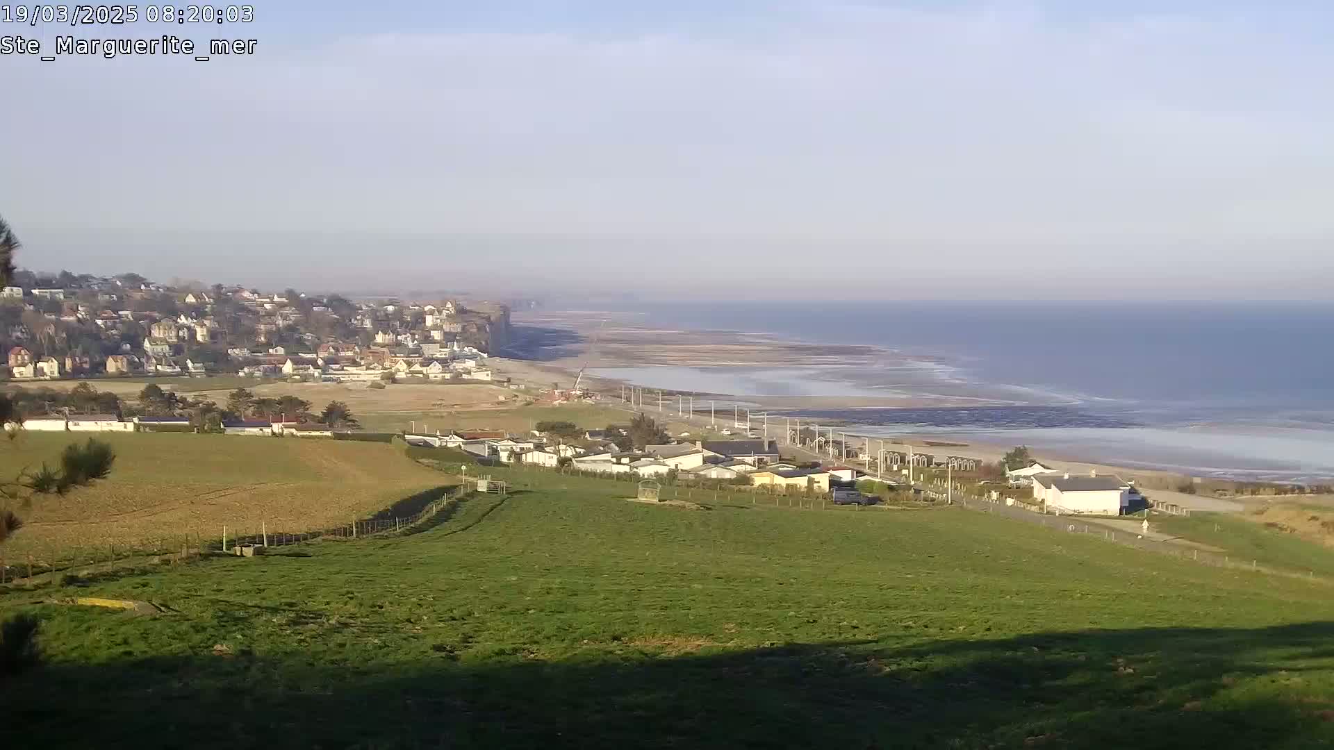 A coastal town nestled on a cliff overlooks a wide expanse of beach and calm ocean under a clear sky.