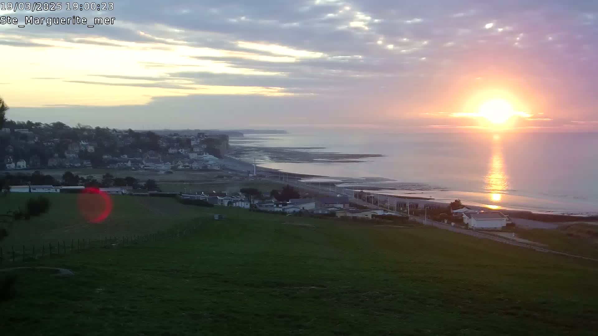 A coastal town sits on a grassy hillside overlooking a calm ocean at sunset under a partly cloudy sky.