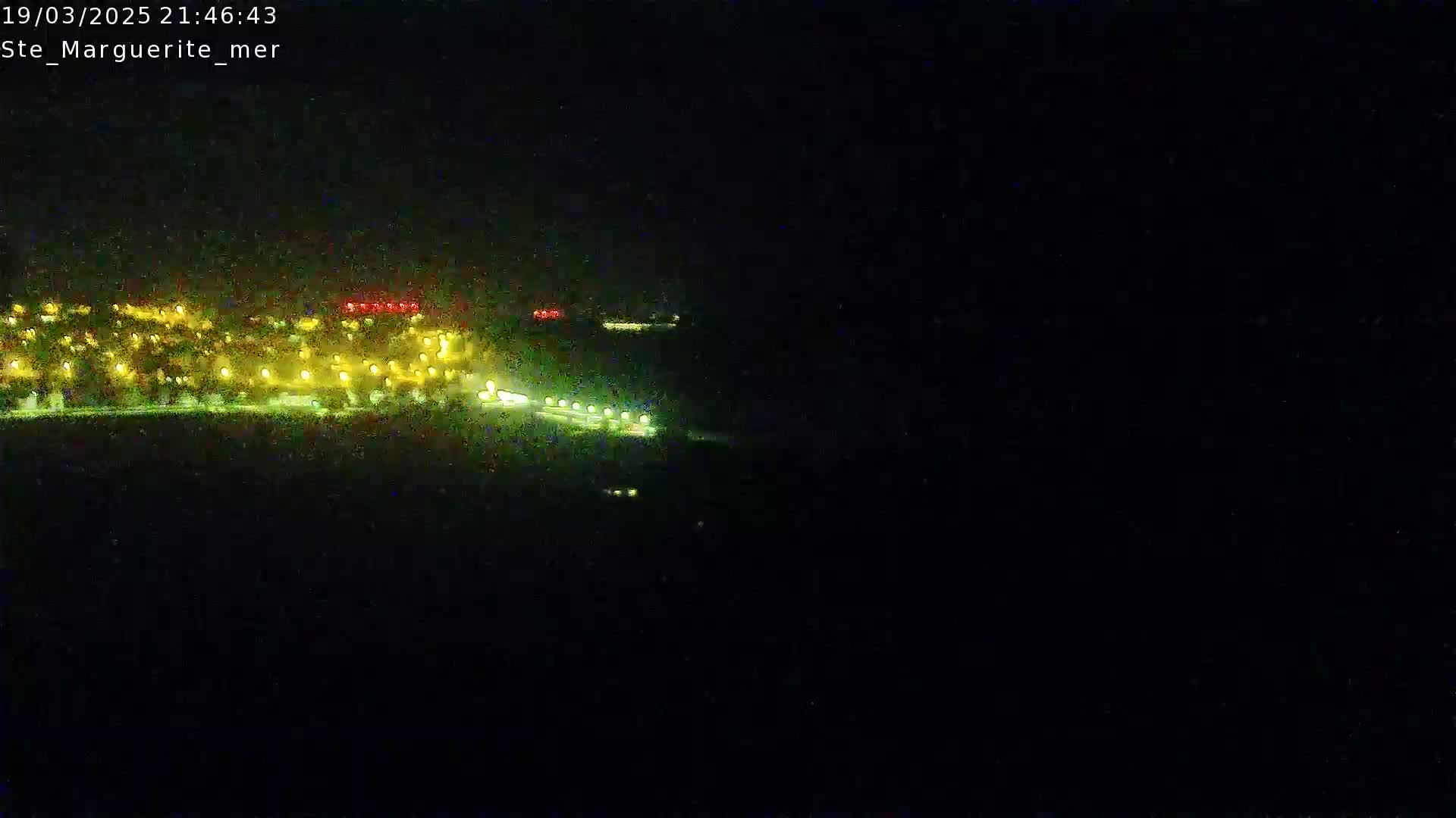 A nighttime long shot shows a brightly lit town or city on the horizon, and a dark body of water extending to the bottom of the frame.