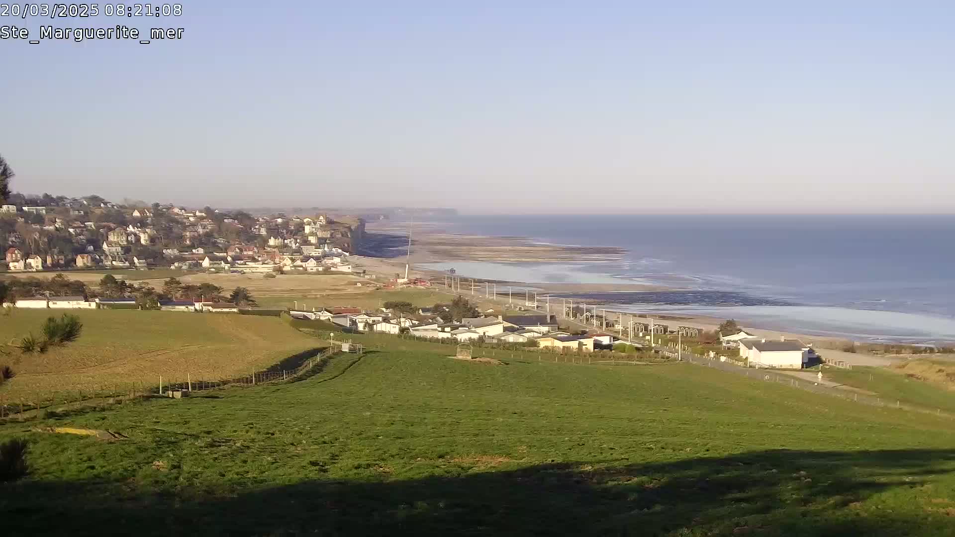 A coastal town sits atop a cliff overlooking a calm ocean, with green fields and a few buildings in the foreground under a clear sky.
