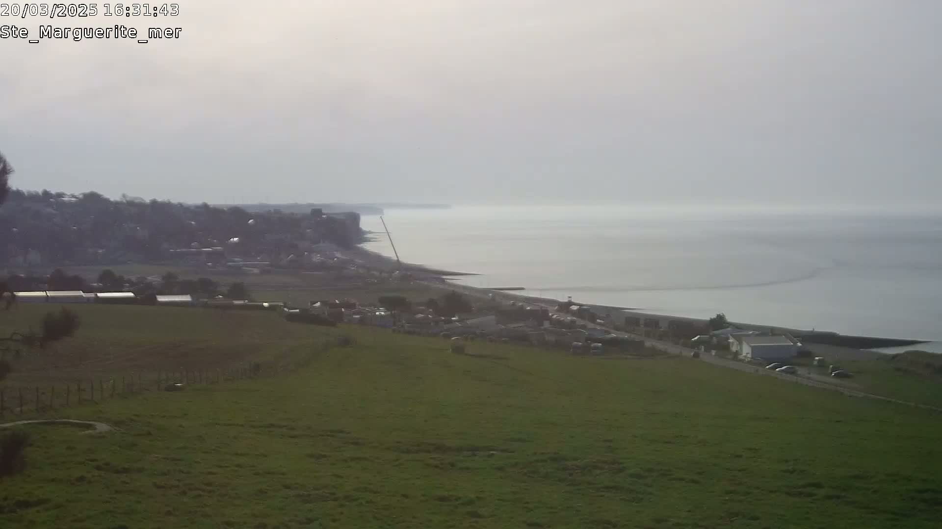 A hazy coastal scene shows a grassy hill overlooking a town, beach, and calm sea under a mostly cloudy sky.