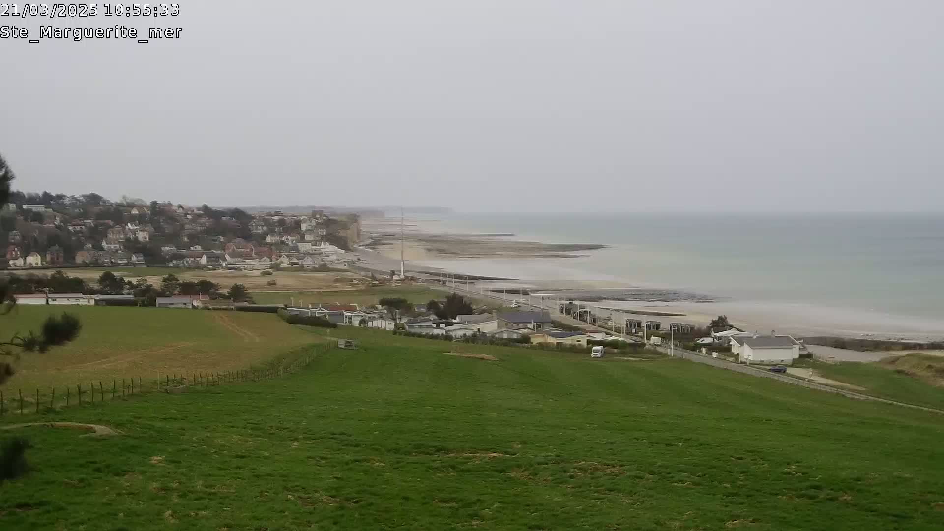 A coastal town overlooks a wide, sandy beach and calm, light-green sea under an overcast sky.