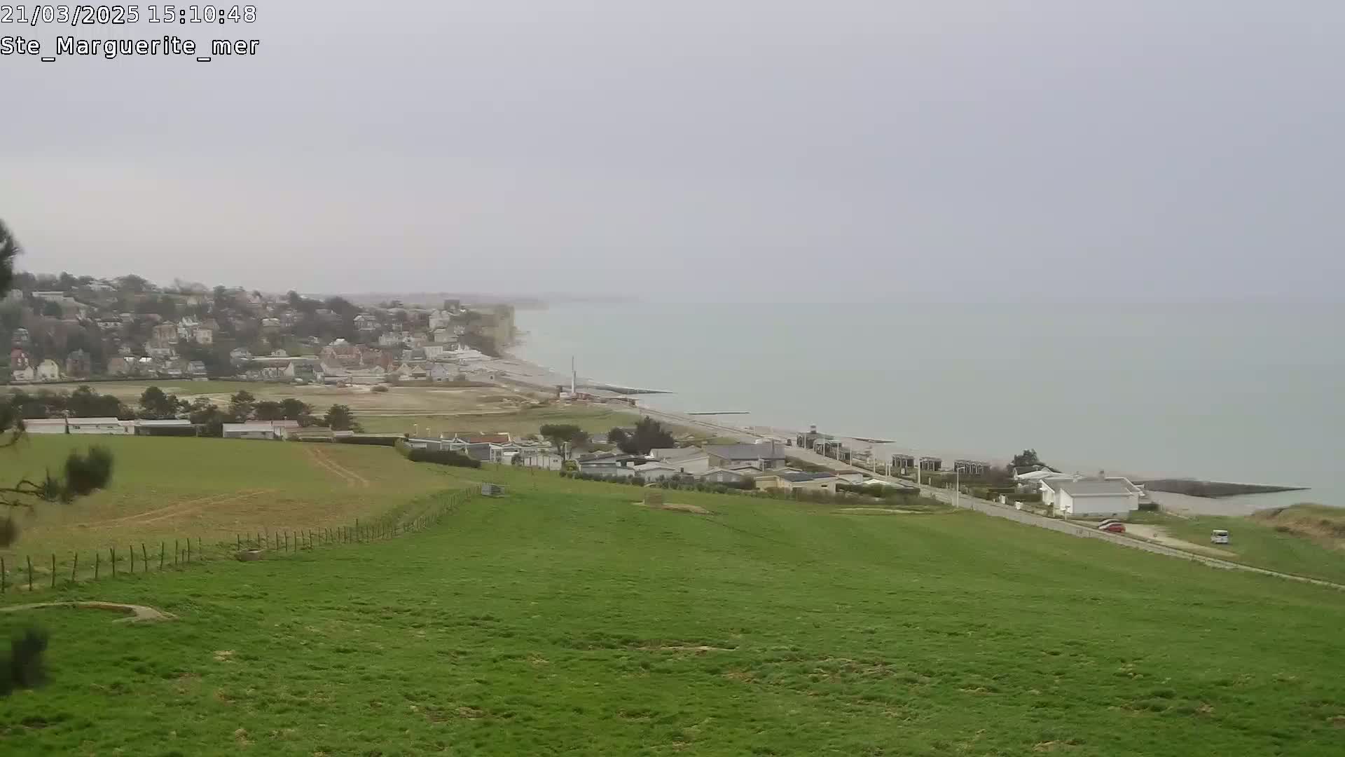 A coastal town sits below a grassy hillside overlooking a calm, gray sea under an overcast sky.