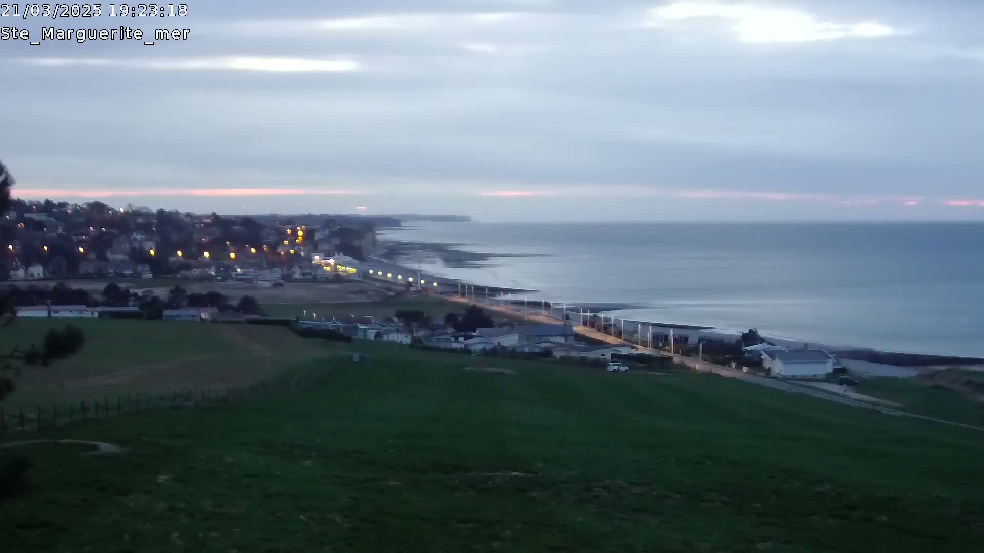 A coastal town at twilight is visible from a hillside overlooking a calm sea under a cloudy sky.