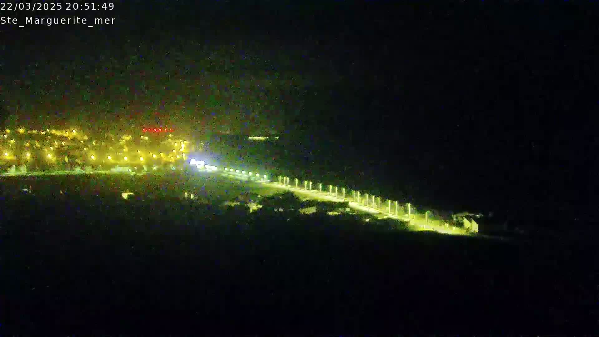 A nighttime, high-angle view shows a brightly lit road curving away from a cluster of buildings toward the darkness.