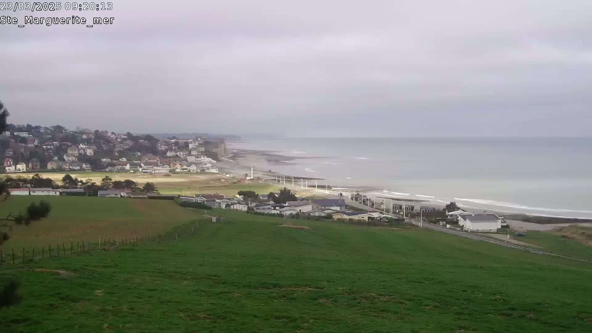 A coastal town sits atop a cliff overlooking a beach with gentle waves under an overcast sky.
