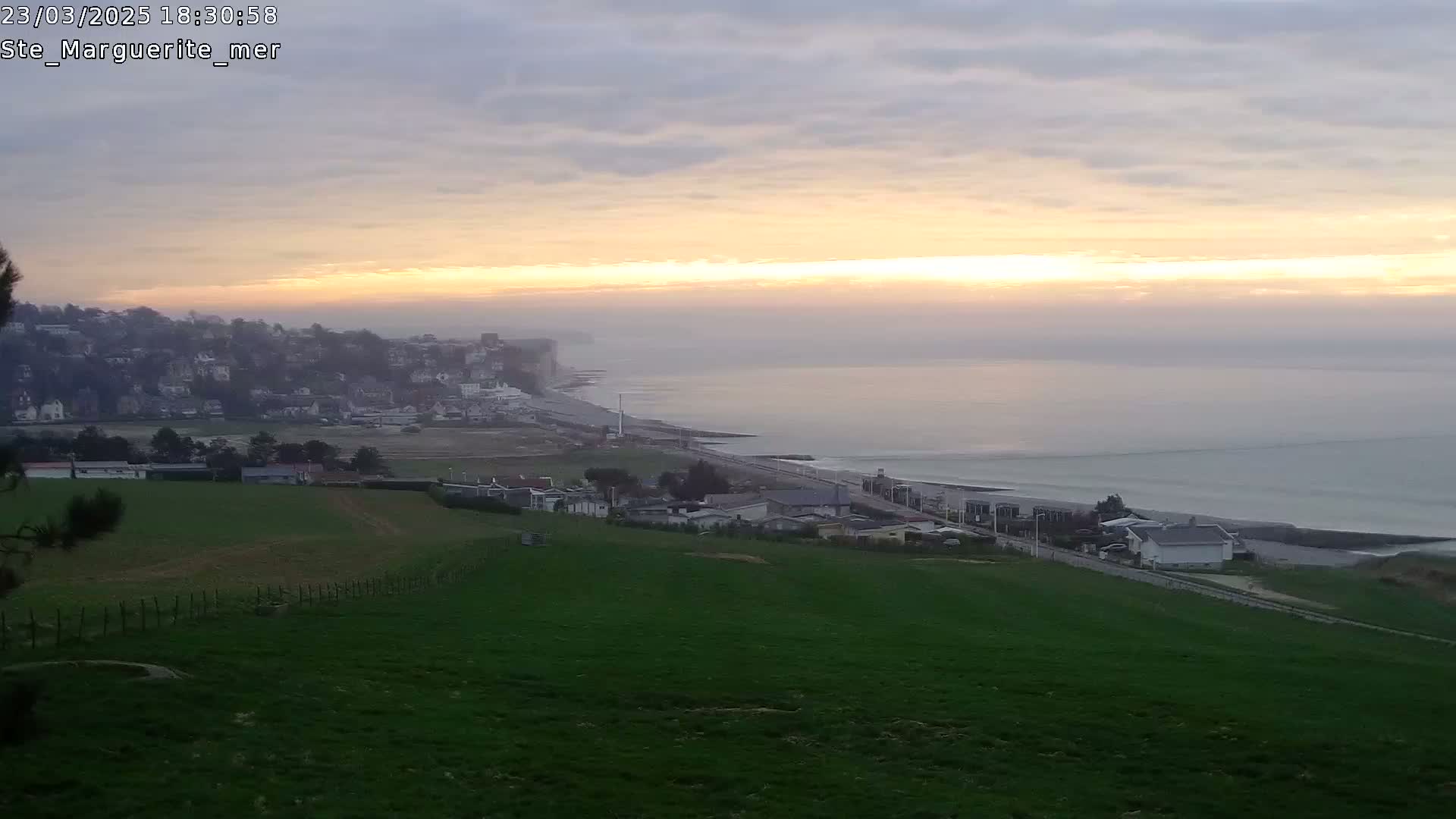 A coastal town sits below a hill of green grass, overlooking a calm ocean at sunset under a hazy sky.