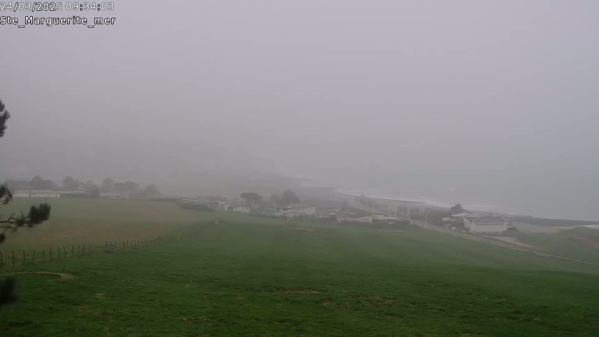 A foggy coastal scene shows a green field sloping down toward a village and a hazy ocean.