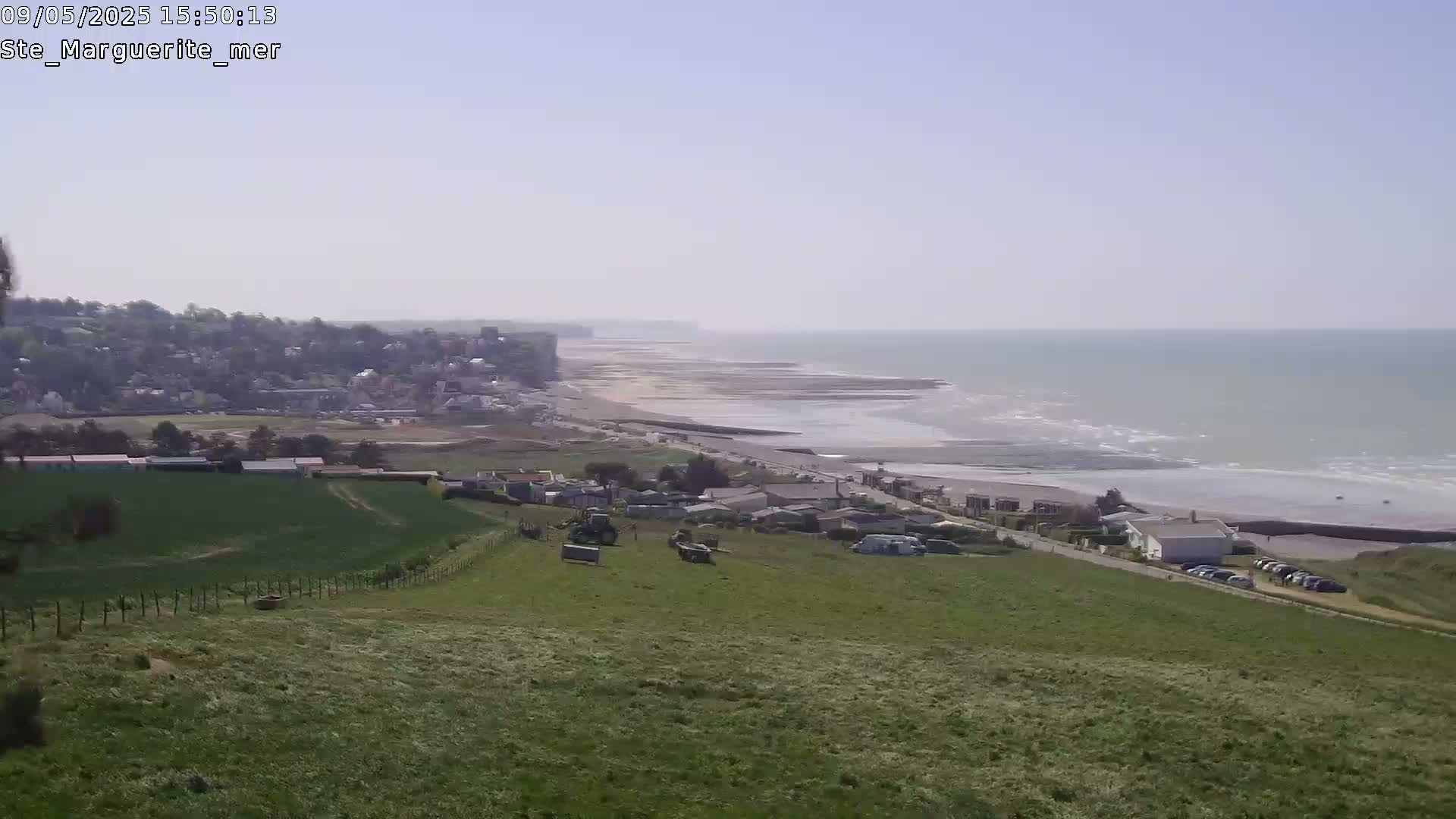 A coastal town sits below a grassy hill overlooking a wide, sandy beach and calm ocean under a clear sky.