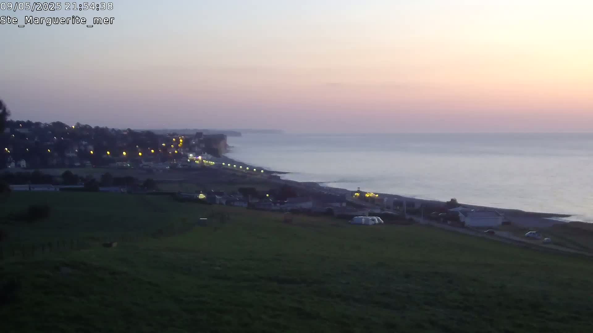 A coastal town at dusk is visible from a hill overlooking a calm sea under a pale pink sky.