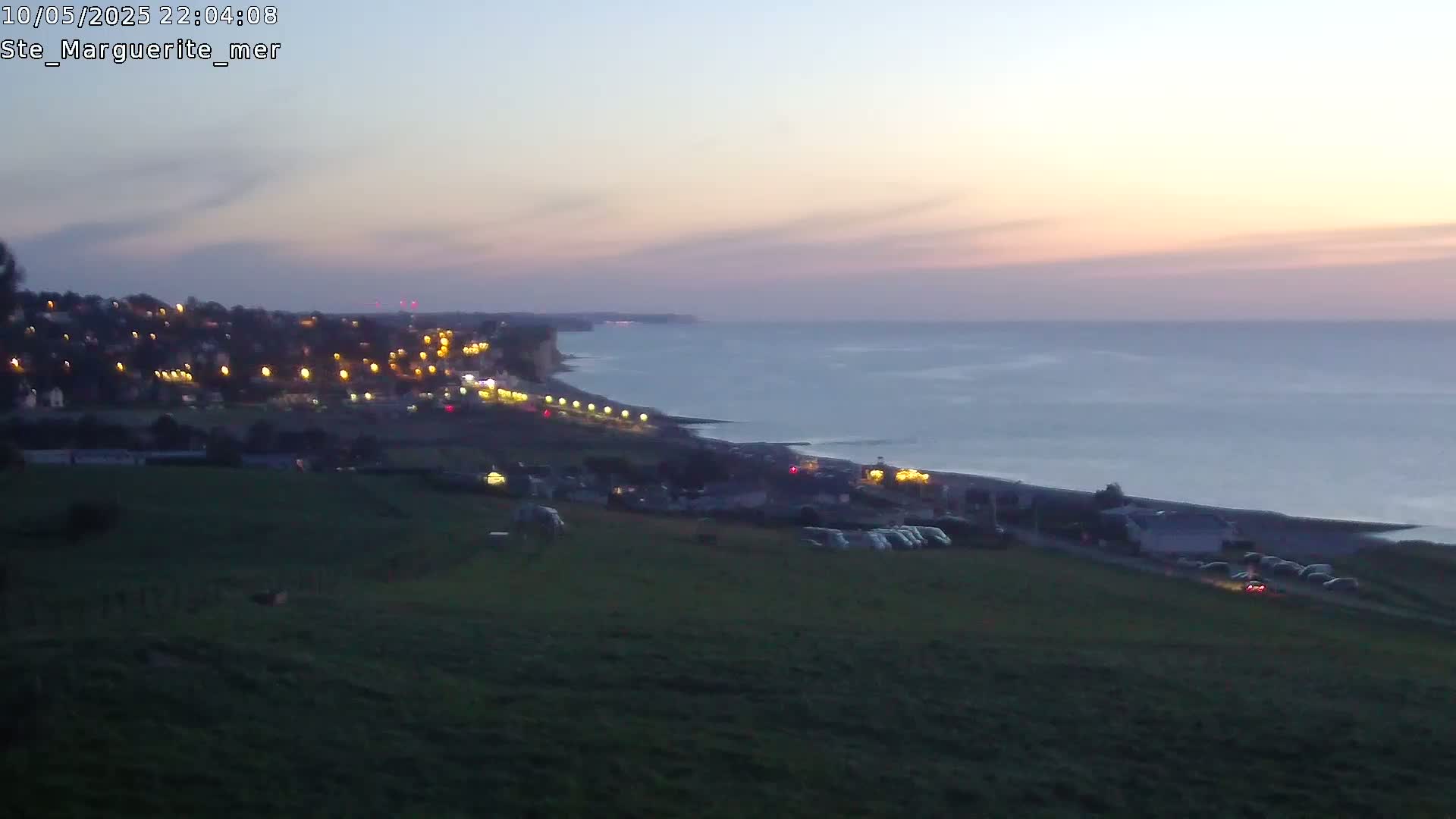 A coastal town at twilight is visible from a hillside, showing a calm sea under a dusky sky.