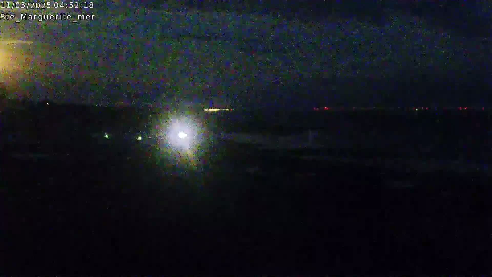 A nighttime, low-light view shows a dark landscape with distant lights, possibly from a town or city, visible on the horizon under a mostly clear sky.