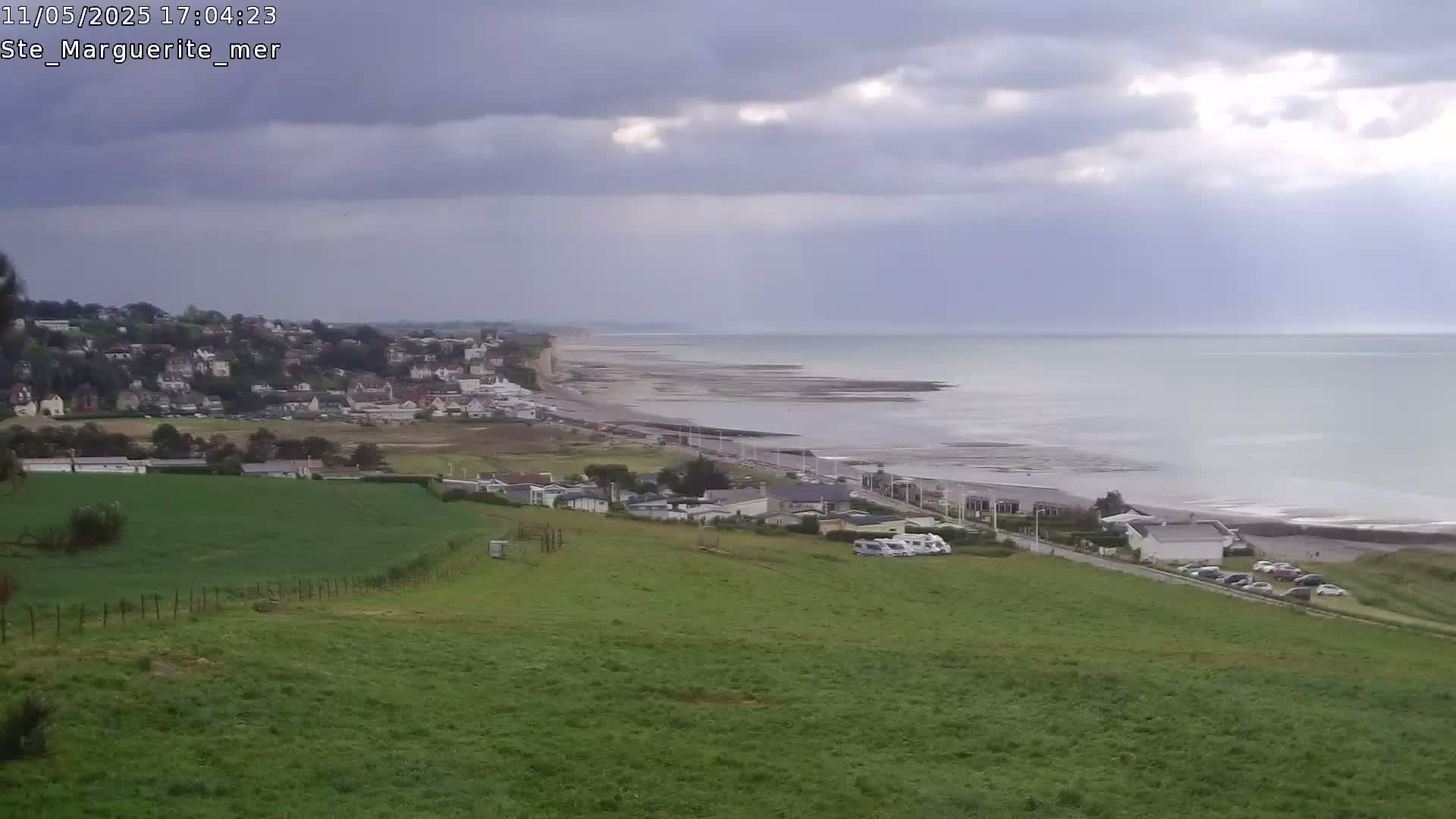 A coastal town overlooks a calm sea under a cloudy sky, with green fields and a few parked vehicles in the foreground.
