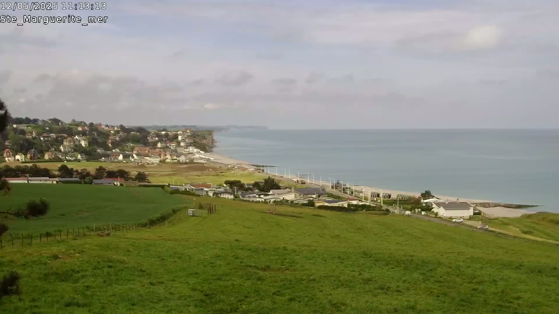A coastal town nestled on a hillside overlooks a calm ocean with a sandy beach under an overcast sky.