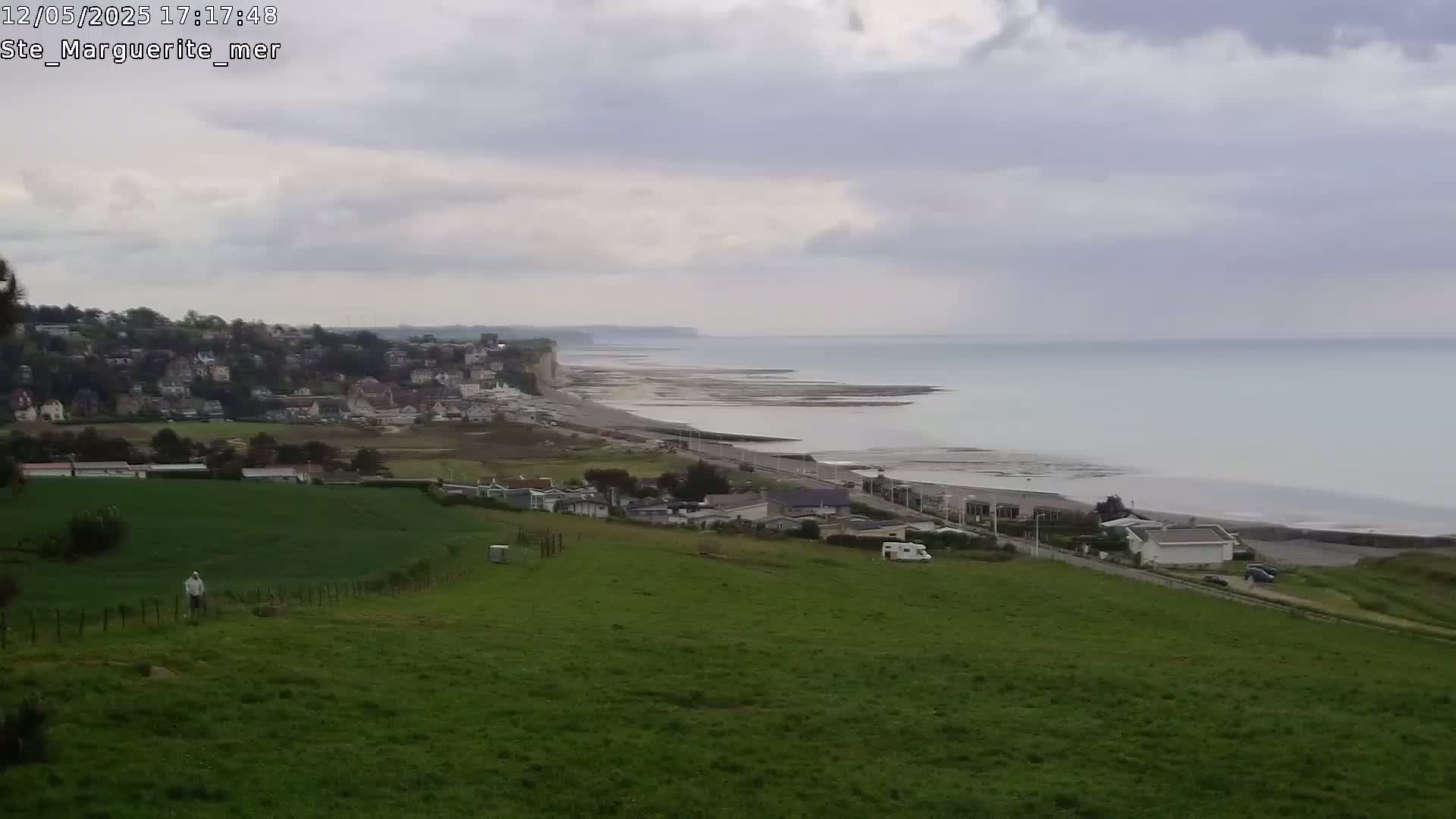 A person walks across a grassy hill overlooking a town, coastline, and calm ocean under an overcast sky.