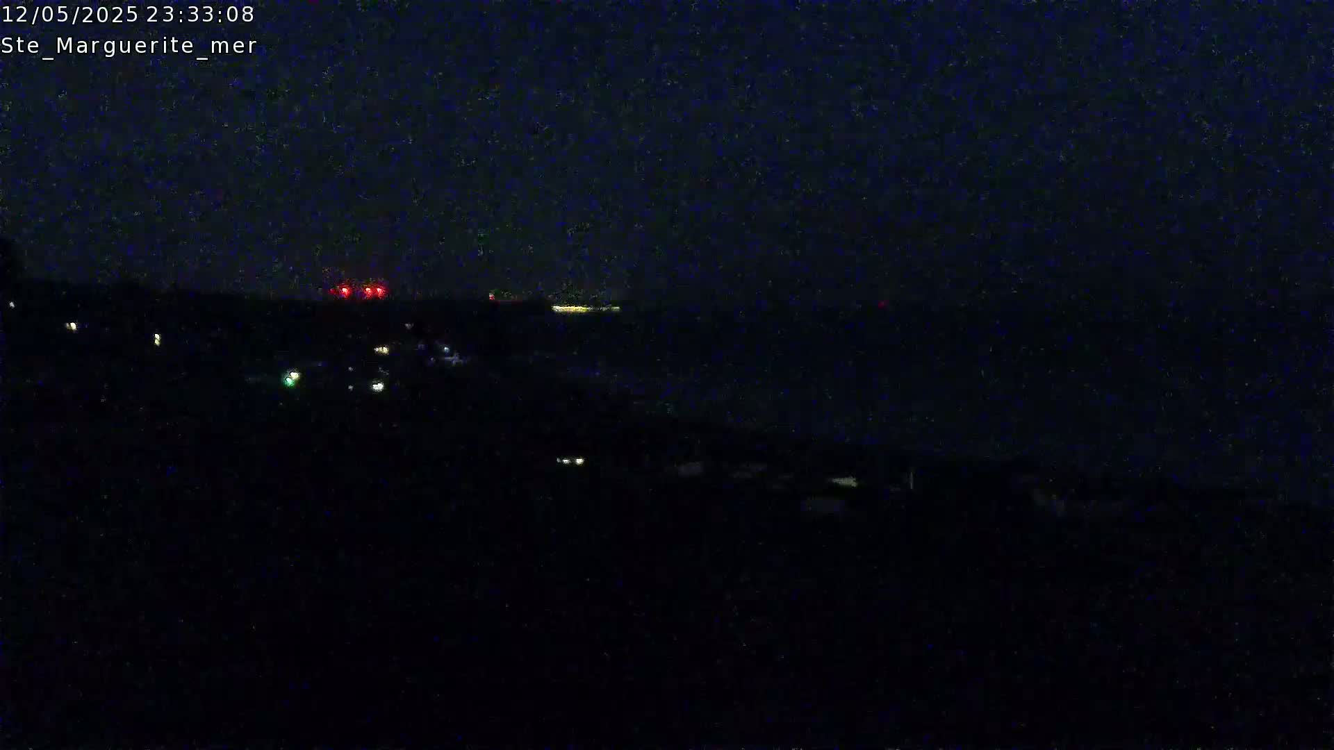 A nighttime, dark view of a coastline with scattered lights visible on land and a faint light source on the water.