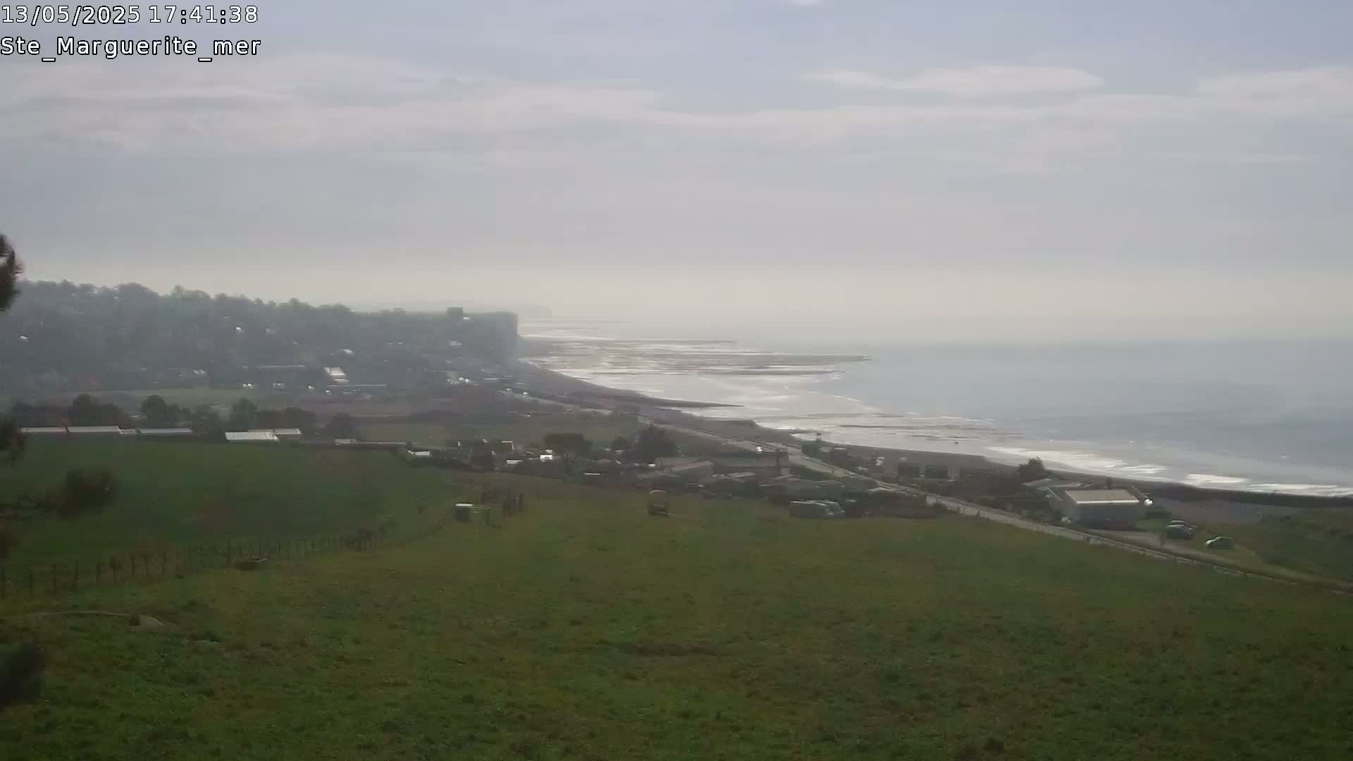A hazy, overcast day reveals a coastal view of a town nestled in green fields overlooking a rocky shoreline and ocean.