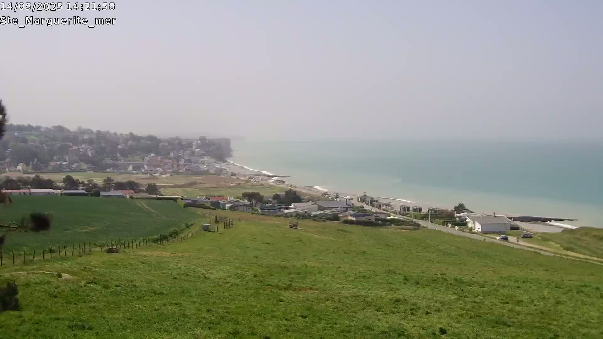 A hazy coastal view shows a green hillside overlooking a town and a calm, teal ocean.