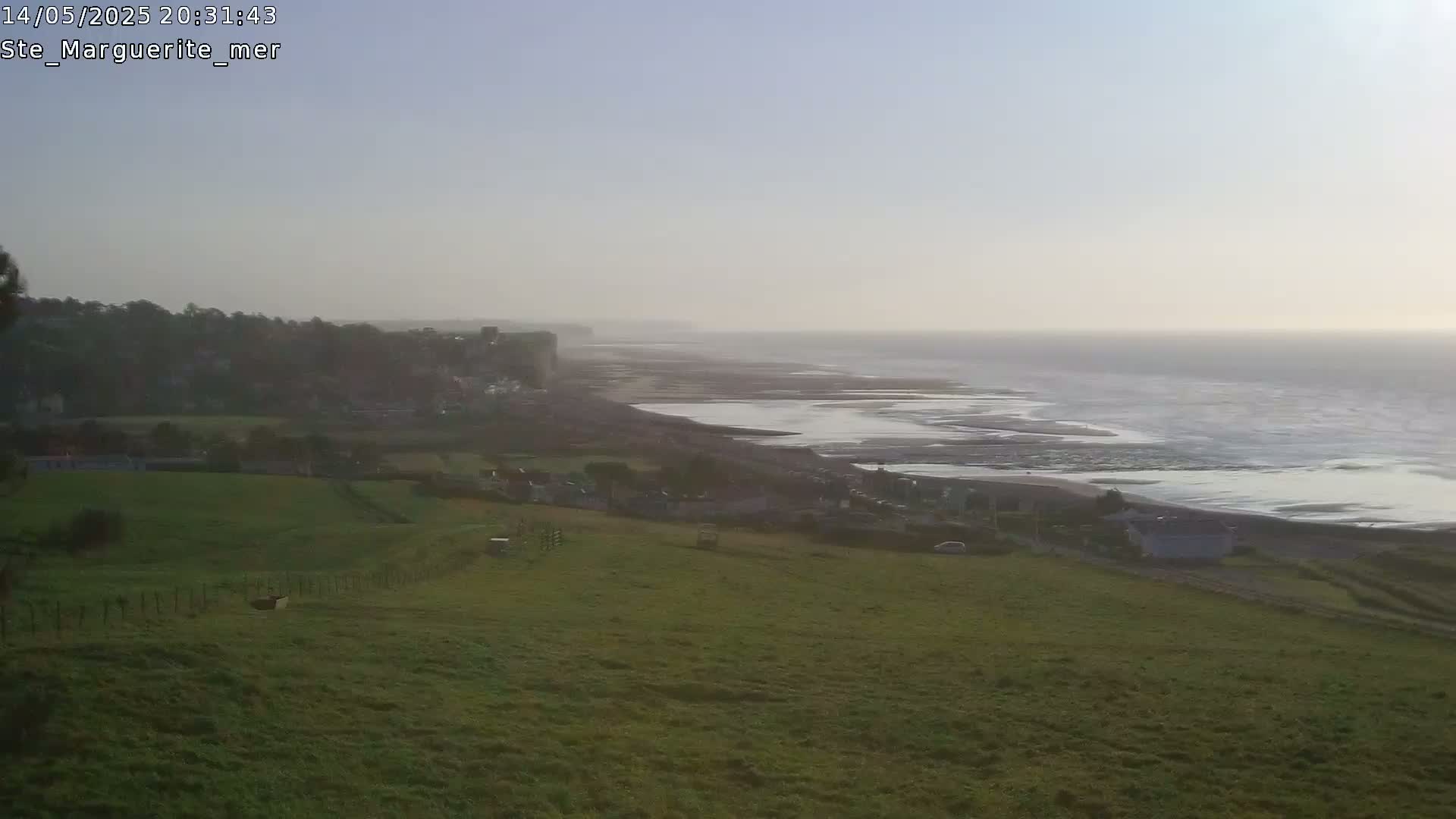 A hazy coastal scene shows a grassy hillside overlooking a village, beach, and ocean under a clear sky.