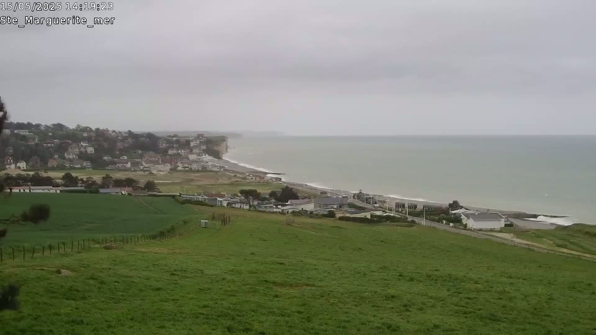 A coastal town nestled below a grassy hill overlooks a gray, overcast ocean with a long, sandy beach.