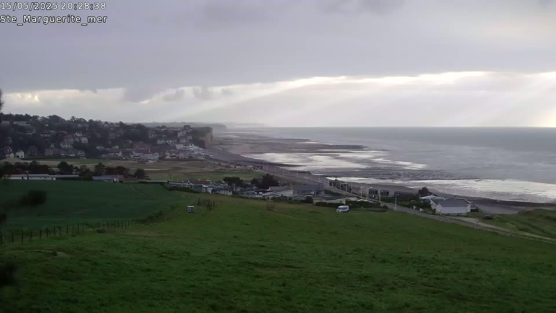 A coastal town sits below a grassy hill overlooking a wide, grey, overcast beach and ocean.