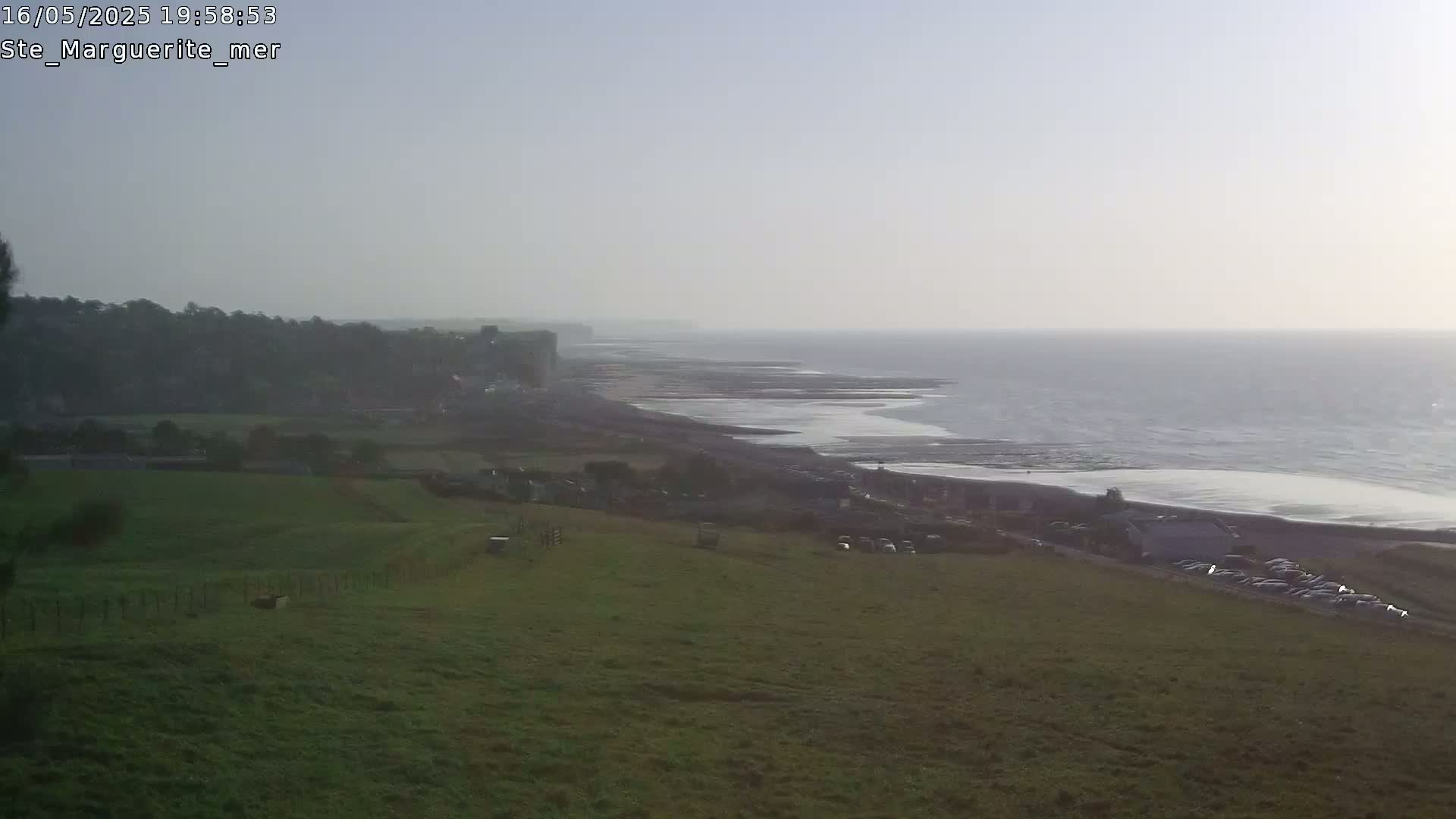 A hazy coastal scene shows a grassy hillside overlooking a beach, ocean, and distant cliffs.