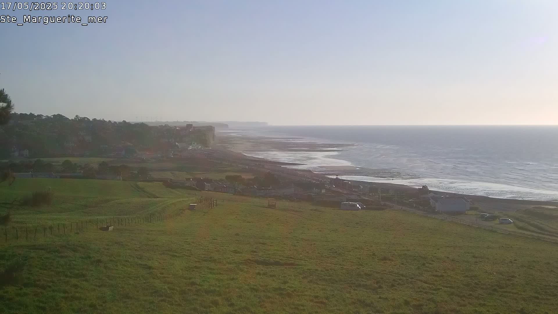 A view from a grassy hill overlooking a calm ocean meeting a long, sandy beach and coastal cliffs under a clear sky.