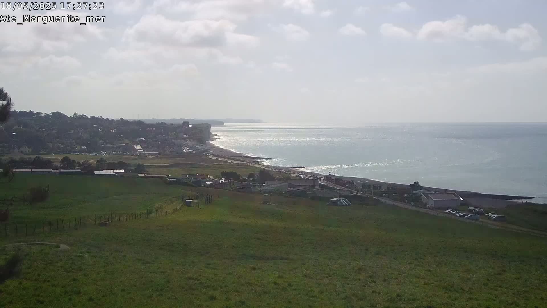 A coastal scene on a partly cloudy day shows a stretch of beach, white cliffs, a town in the distance, and a foreground of grassy fields.
