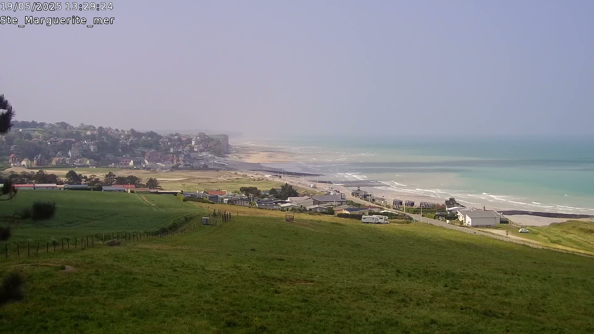 A hazy coastal scene shows a town nestled on a cliff overlooking a sandy beach and turquoise ocean with gentle waves.