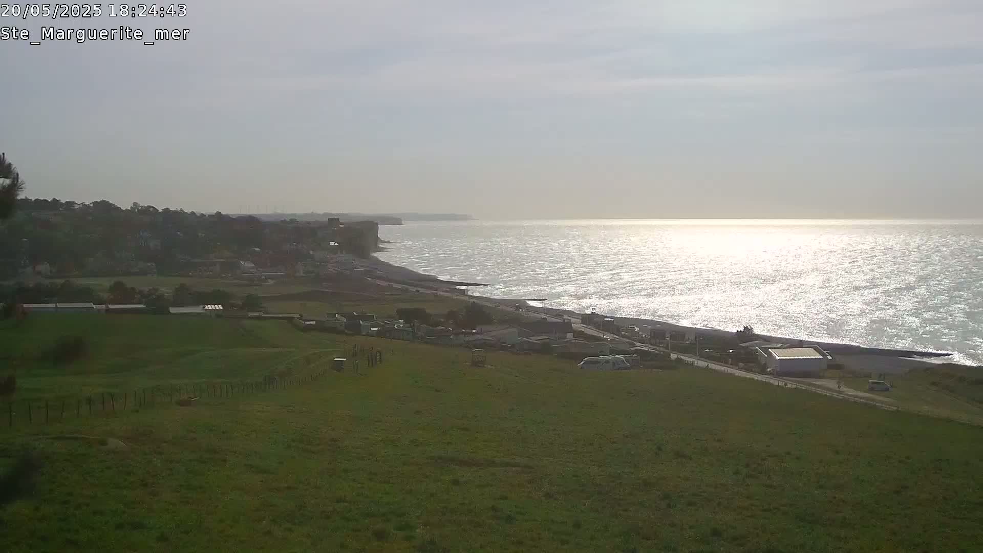 A grassy hillside overlooks a coastline with white cliffs, a village, and a shimmering sea under a mostly cloudy sky.
