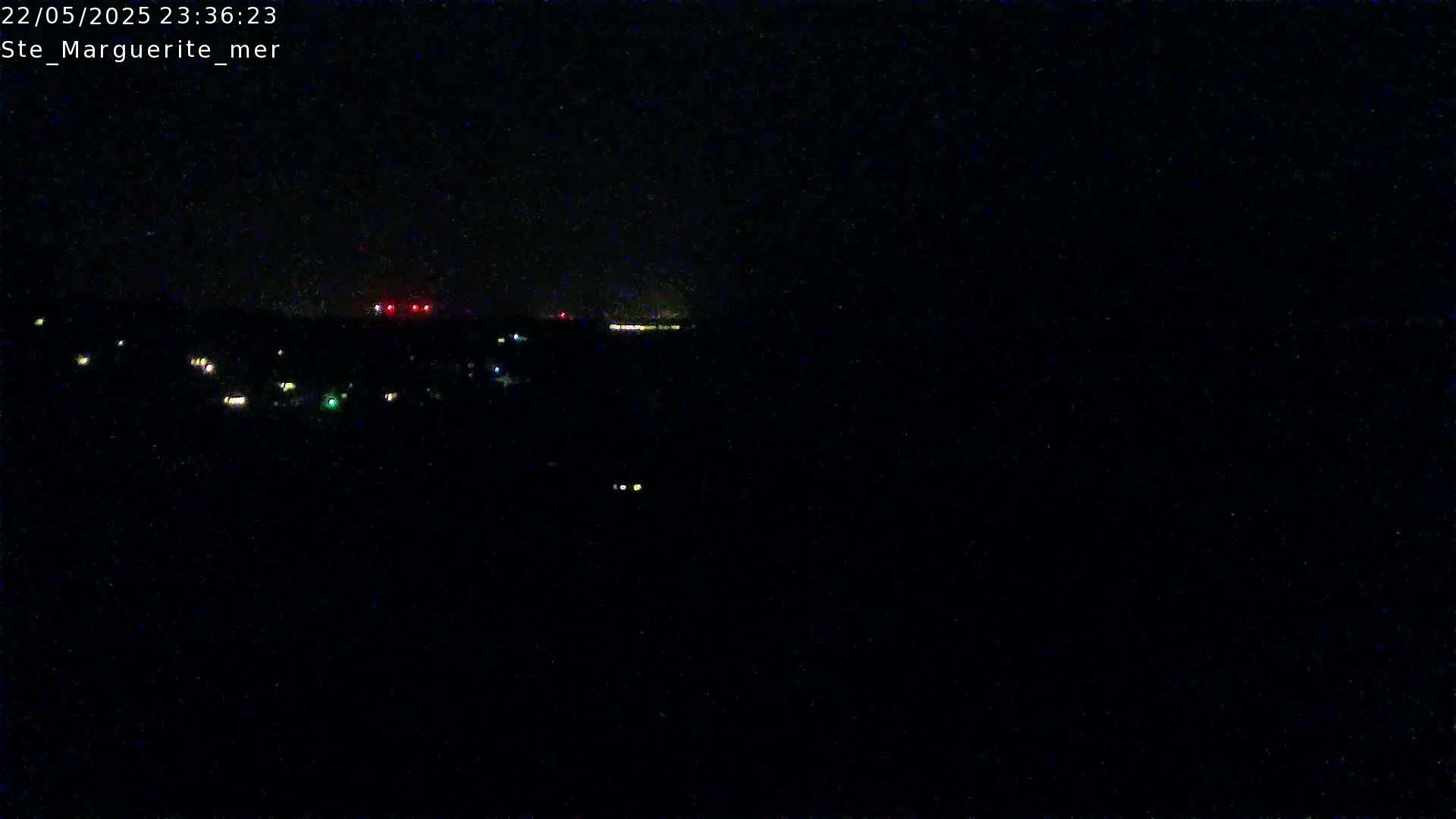 A nighttime long shot shows a dark landscape with scattered, faint lights and a brighter light source in the distance under a clear, dark sky.
