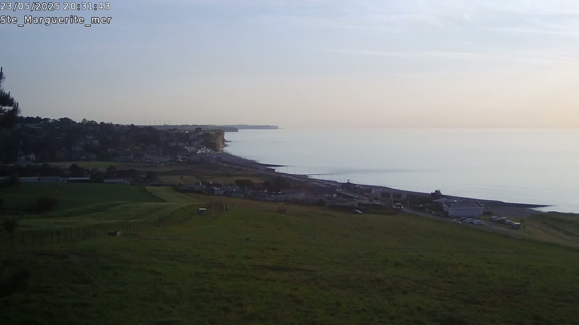 A high-angle view shows a coastal town nestled between a grassy hillside and a calm, pale-blue ocean meeting a dark cliff in the soft light of dawn or dusk.