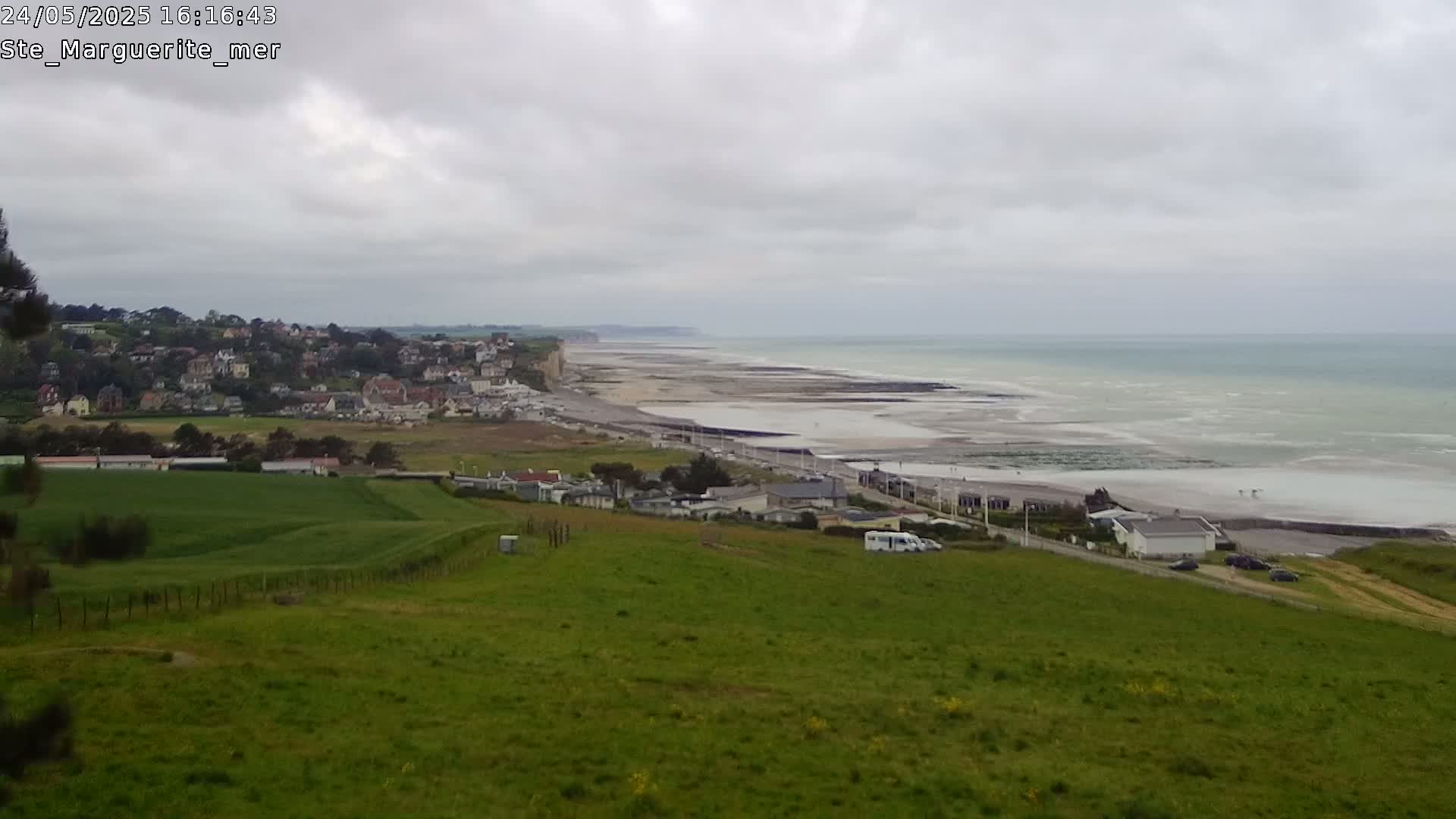 A coastal town sits below a grassy hill overlooking a wide, sandy beach and calm ocean under an overcast sky.