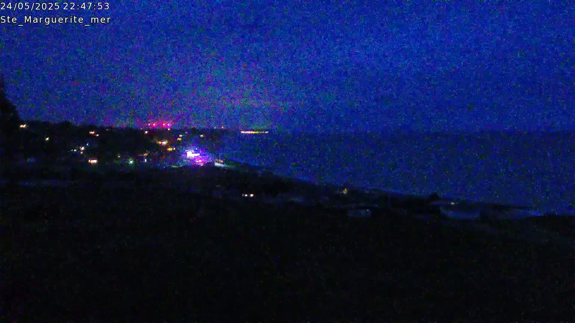A dark, nighttime coastal scene shows a dark body of water under a deep blue twilight sky, with scattered lights visible on a hillside in the foreground.