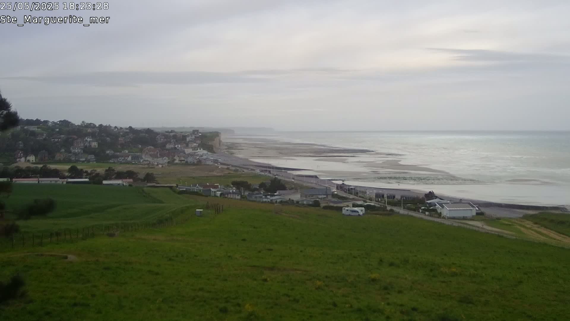 A coastal town sits below a hillside of green grass overlooking a wide, light-grey ocean under an overcast sky.