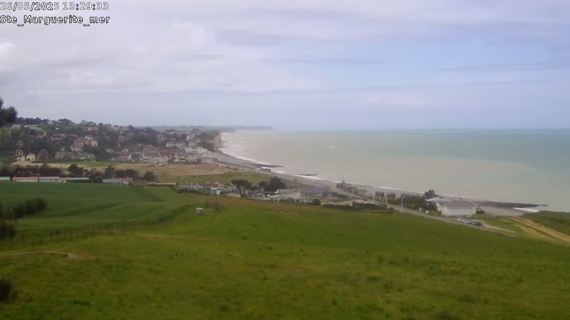 A coastal town nestled beside a grey, overcast ocean, with rolling green hills in the foreground.