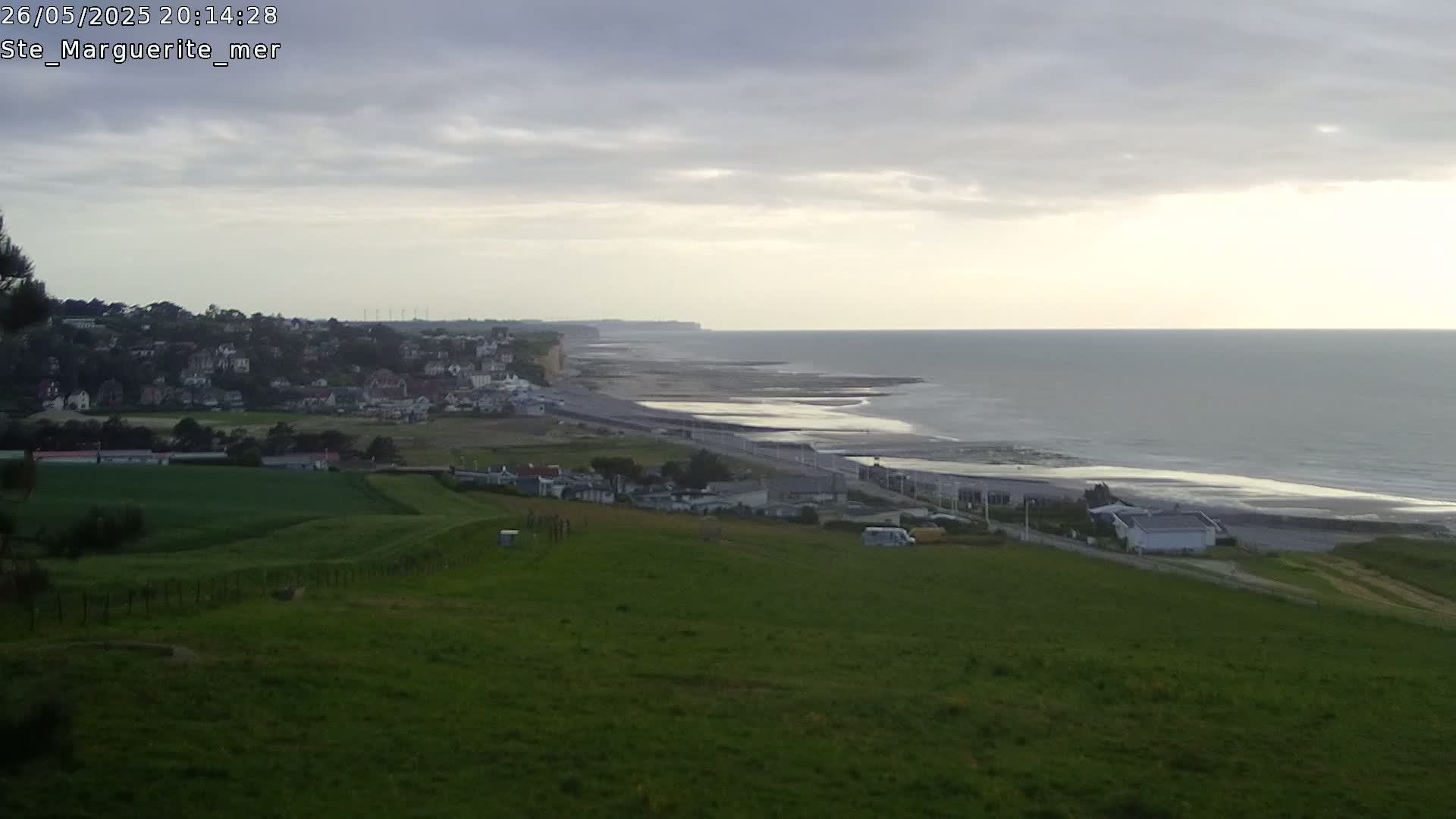 A coastal town sits below a verdant hillside overlooking a calm ocean under a cloudy sky.