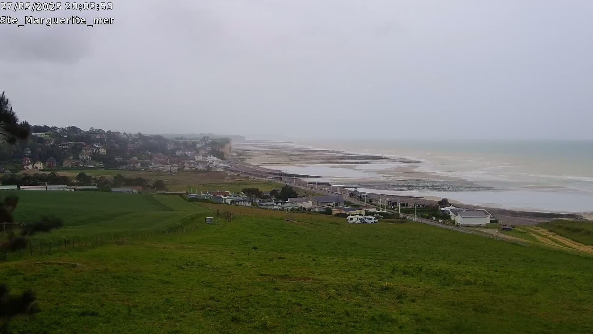 A coastal town sits atop a cliff overlooking a wide, sandy beach and calm, gray sea under an overcast sky.