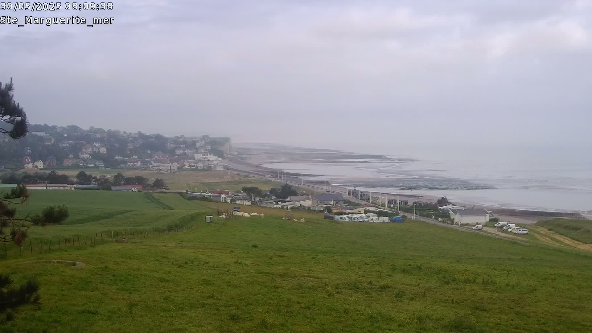 A hazy coastal view shows a town nestled against a cliff overlooking a wide expanse of ocean, with green fields and several parked vehicles in the foreground.