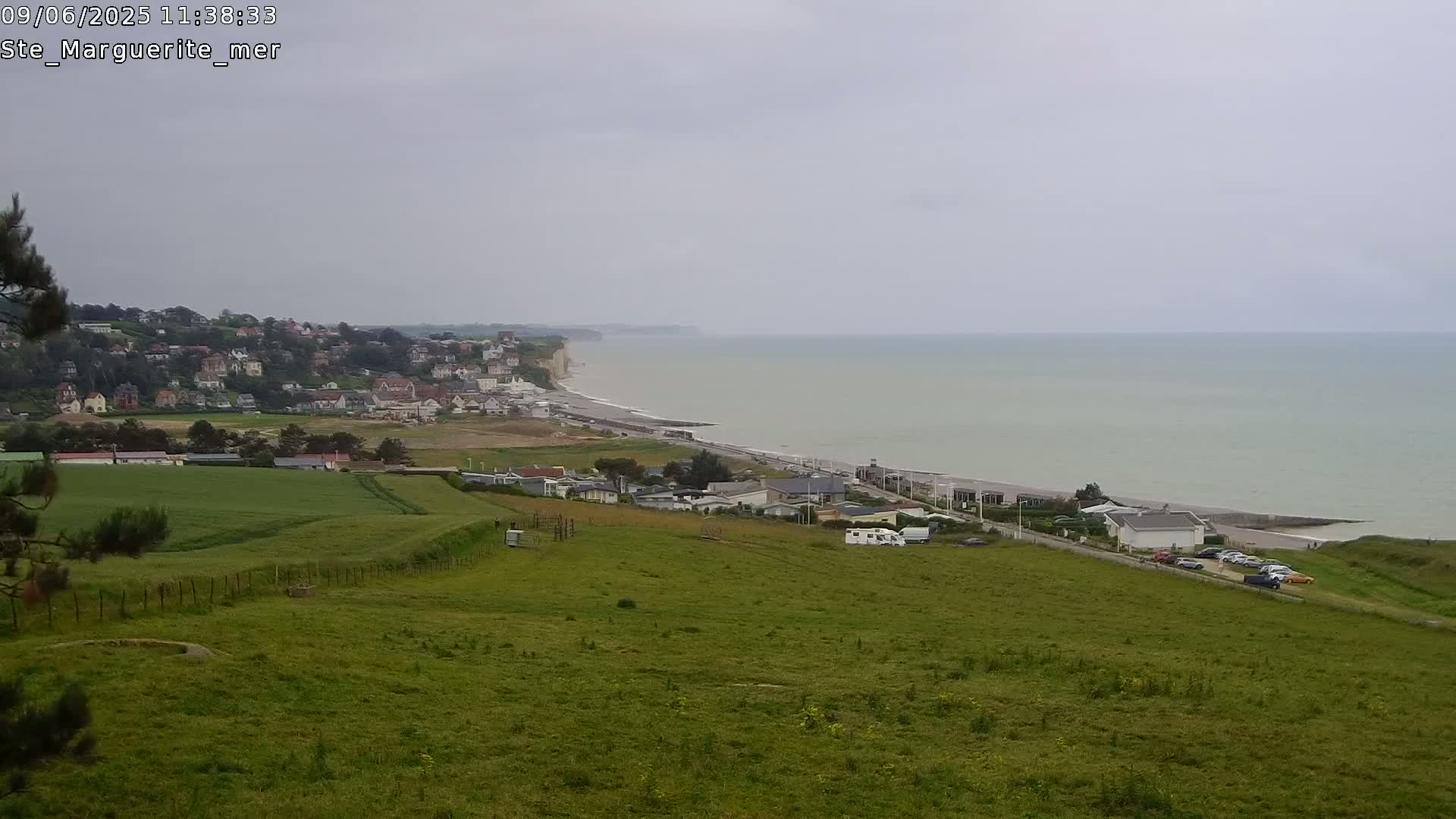 A coastal town sits below a grassy hill overlooking a calm, gray sea and white cliffs under an overcast sky.