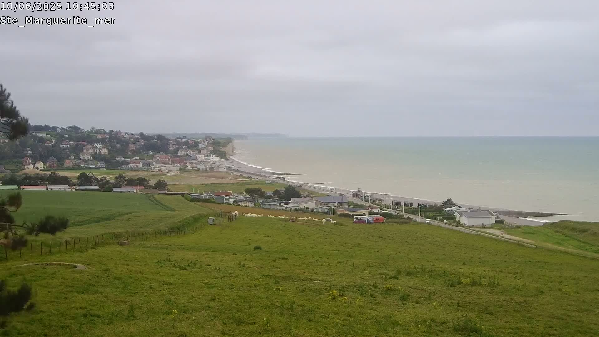 A coastal town sits below a grassy hill overlooking a calm, gray sea meeting a sandy beach.