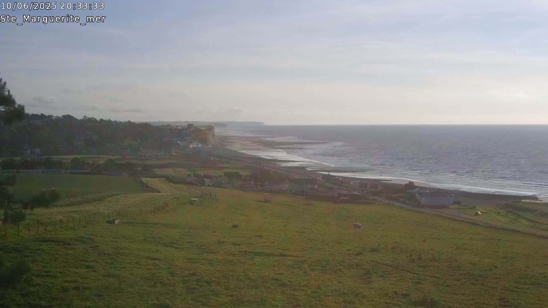A grassy hillside overlooks a town, a beach, and a calm ocean under a hazy sky.