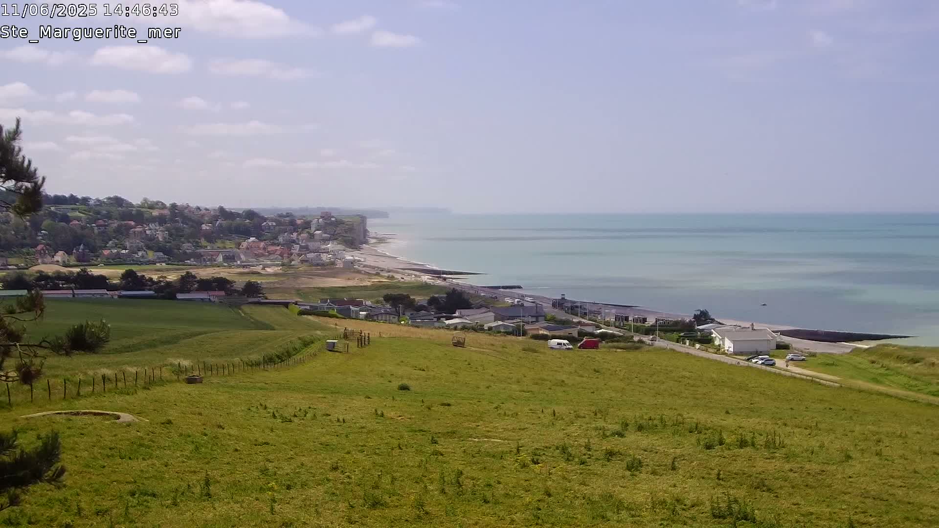 A coastal town sits below a grassy hillside overlooking a calm sea and a long, sandy beach stretching along a cliffside.