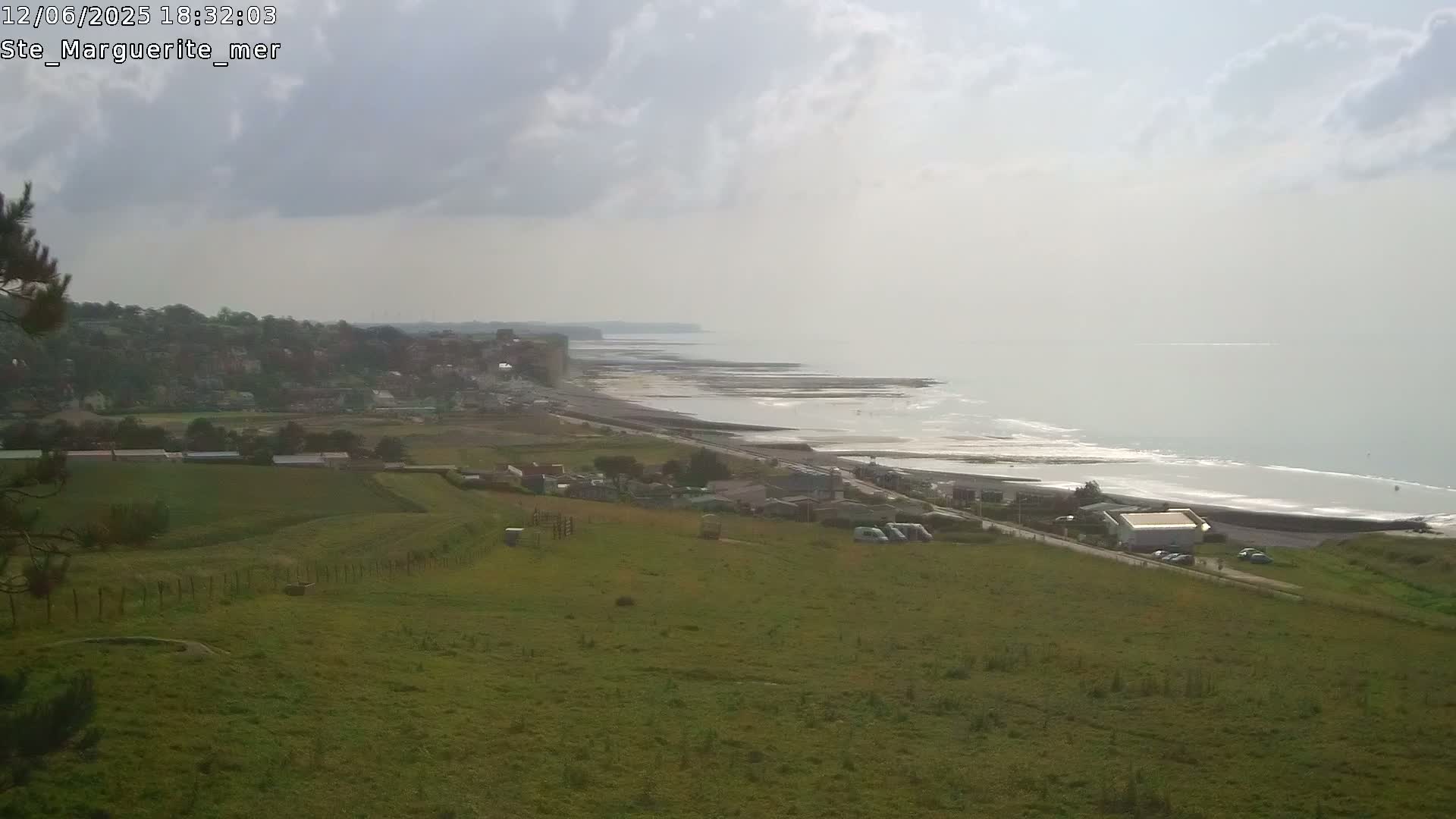 A coastal scene under a cloudy sky shows a grassy hillside overlooking a town, a beach, and a body of water with whitecaps.