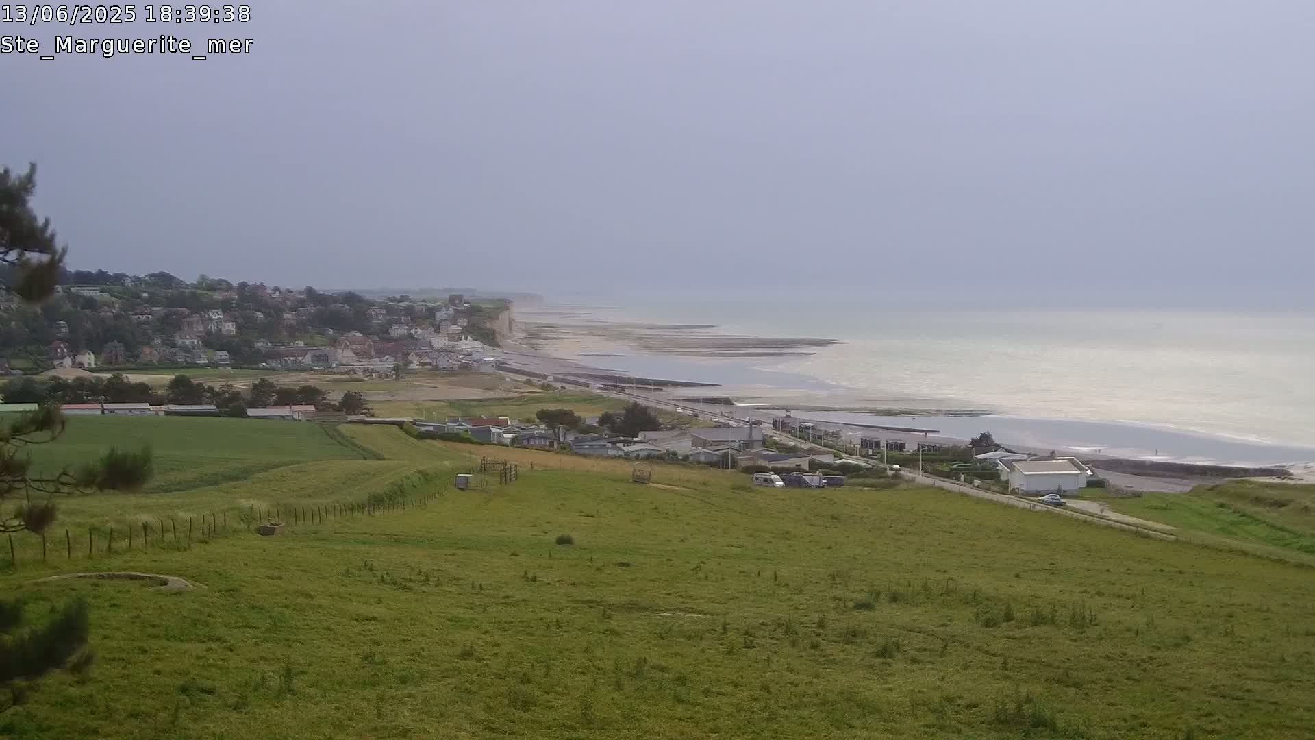 A coastal town sits below a grassy hillside overlooking a calm sea under an overcast sky.
