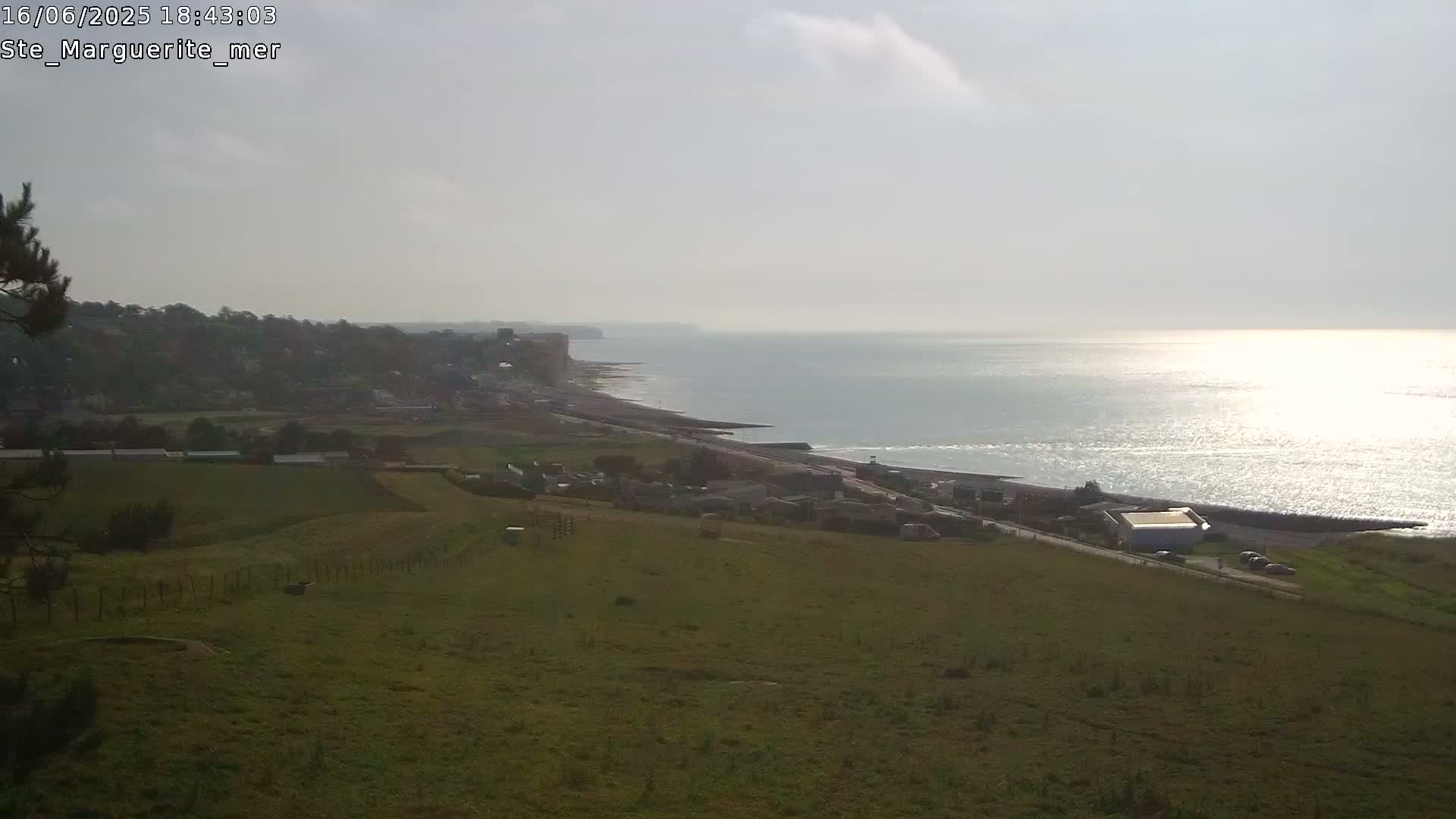 A hilly, grassy landscape overlooks a calm sea reflecting sunlight, with a distant coastline featuring buildings and cliffs under a hazy sky.