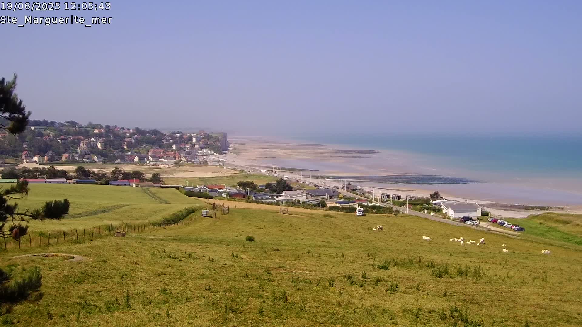 A high-angle view shows a coastal town nestled against a cliff overlooking a wide sandy beach and calm ocean under a clear blue sky.