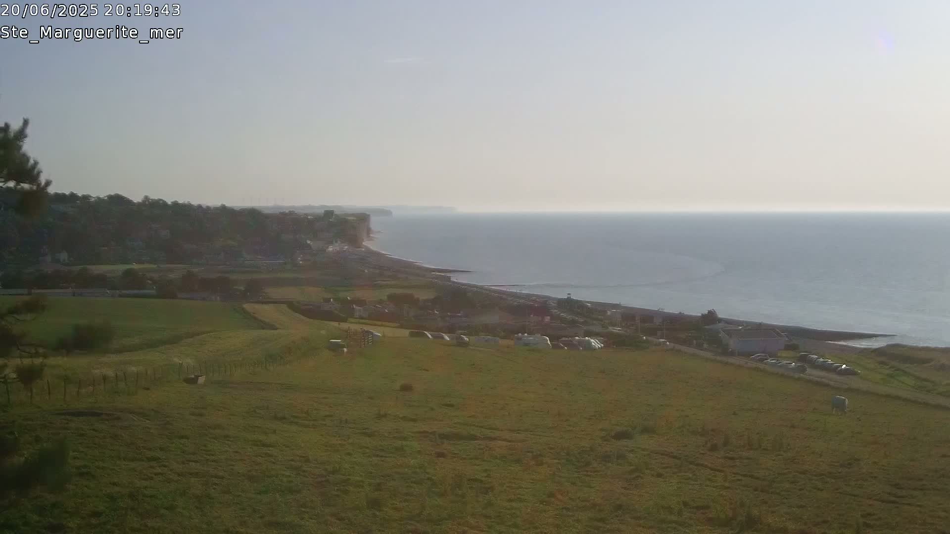 A vast expanse of calm ocean meets a gently sloping coastline dotted with buildings and caravans, overlooking a field under a hazy sky.